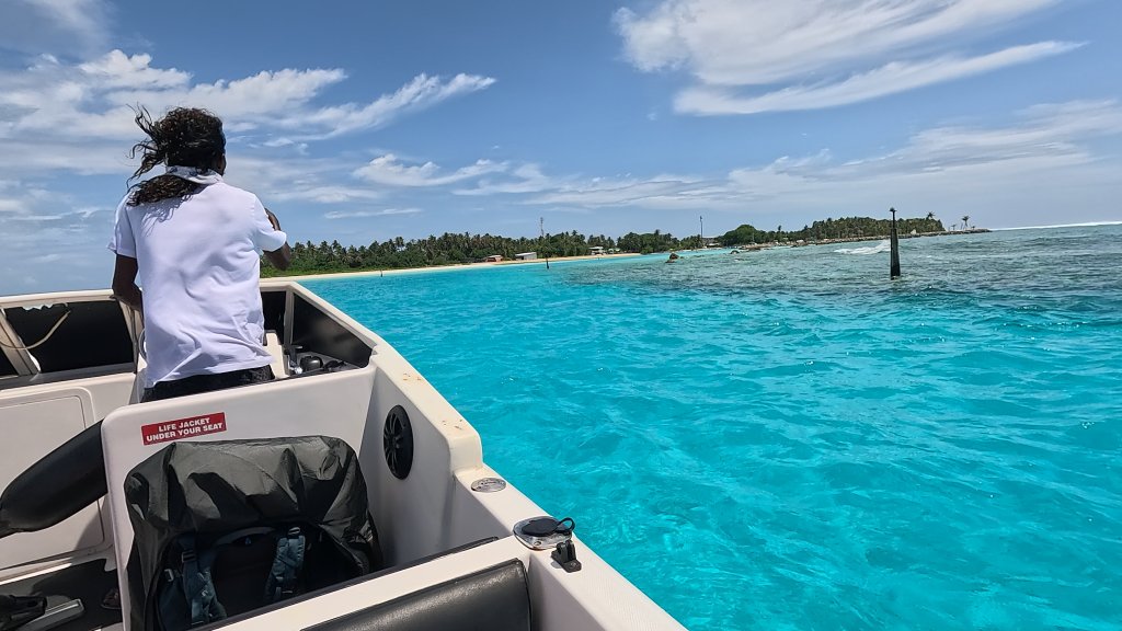 a boat enters the harbour at fulhadhoo over bright turquoise waterm with fulhadhoo island in the distance